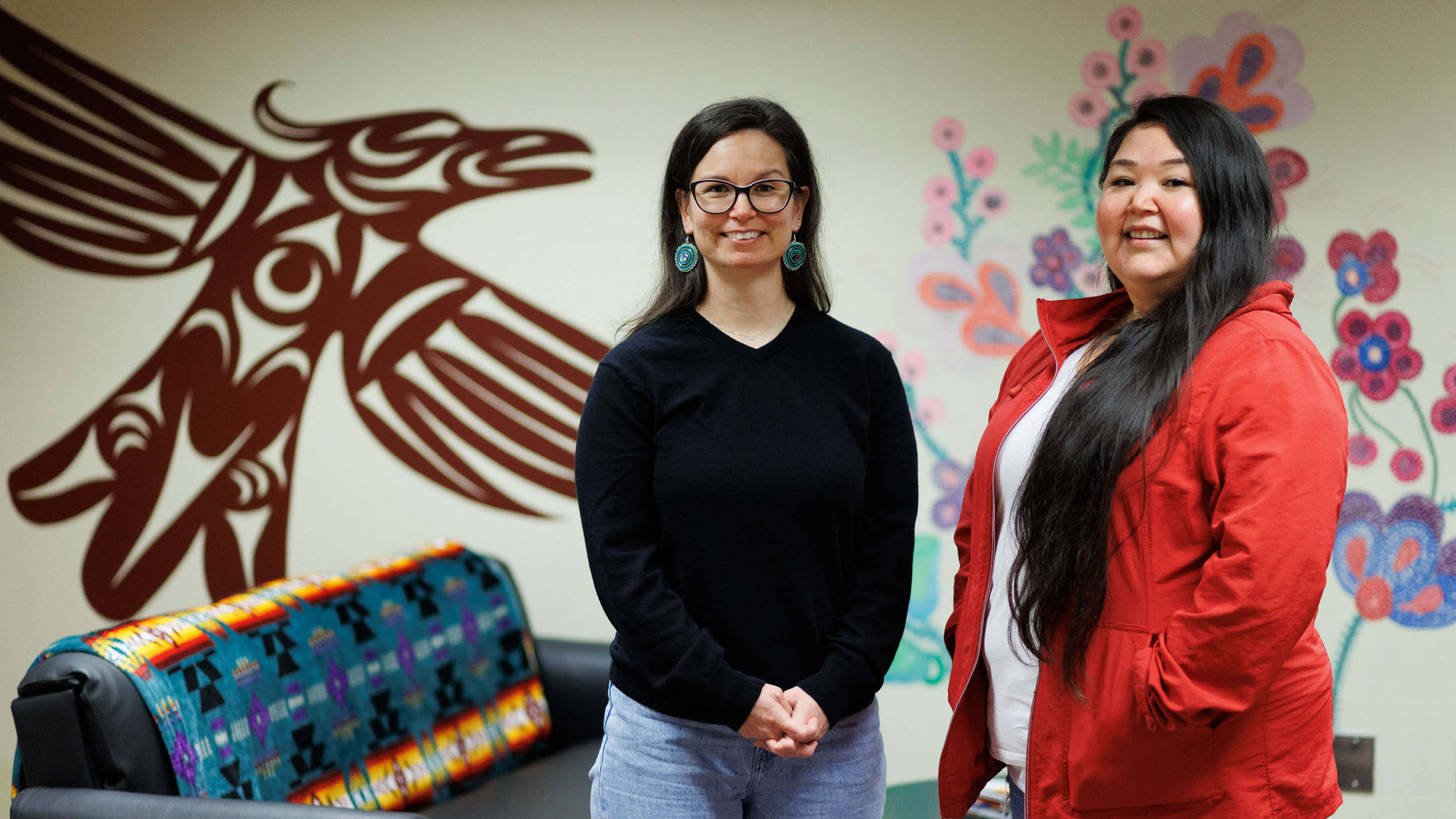 Christina Tsuil-menak and Kyla Elliott, fourth year UVic nursing students. A wall featuring Indigenous artwork of a bird and flowers is behind them.