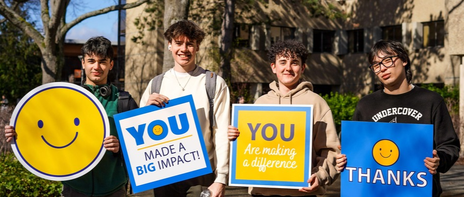 4 students hold signs saying "thanks", "you are making a difference" "you made a big impact" and a yellow smiley face while smiling at the camera. 