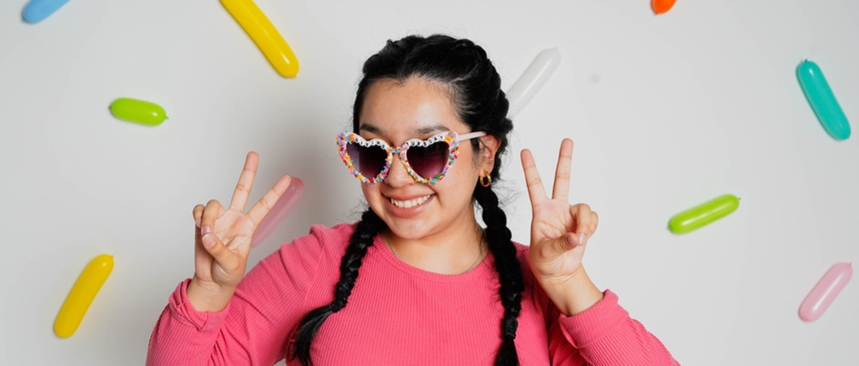 girl wearing pink sweater and sprinkle glasses that say "giving tuesday" smiles at camera while holding two peace signs 
