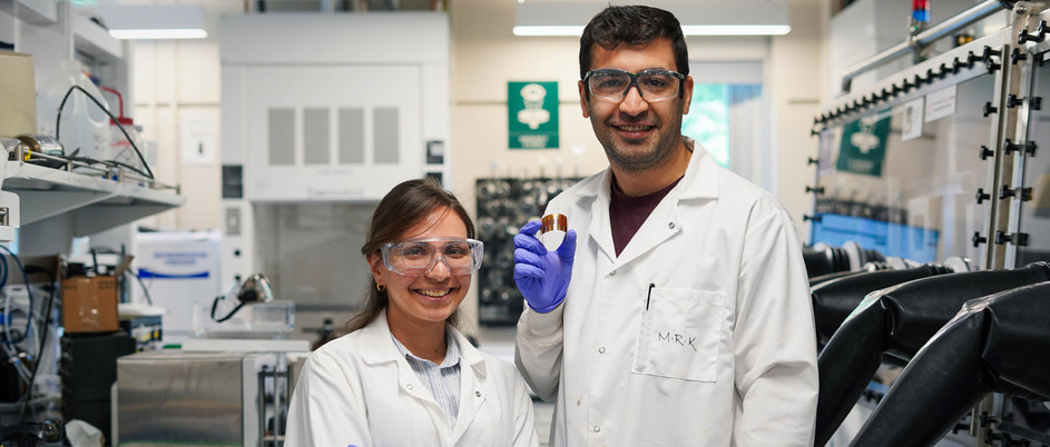 Parinaz and her husband and colleague, Reza, stand in a lab wearing lab coats. Reza is holding a flexible solar cell. 