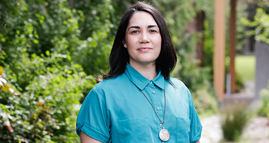 A woman standing in front of greenery looks directly into the camera