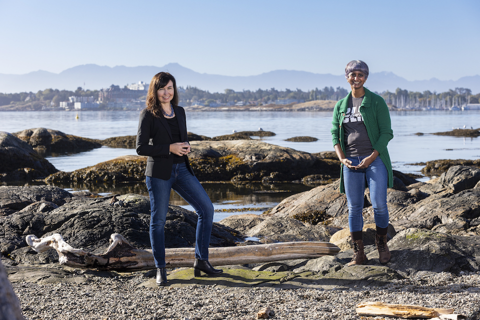 Julia Baum and Hansi Singh standing on rocks by the edge of the water, with building in the background