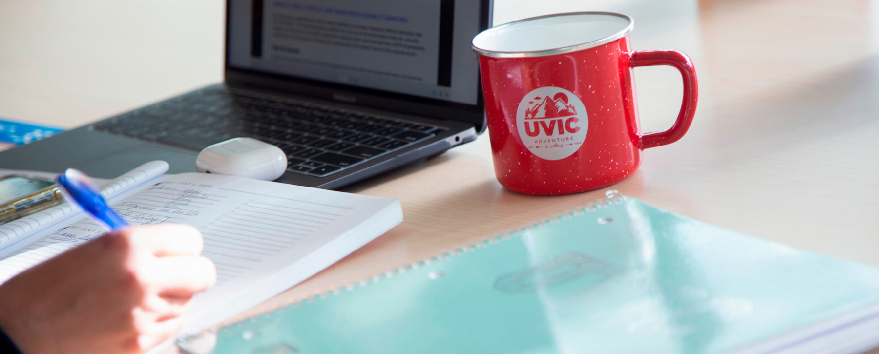 Person working at a desk with a notepad, laptop and UVic mug