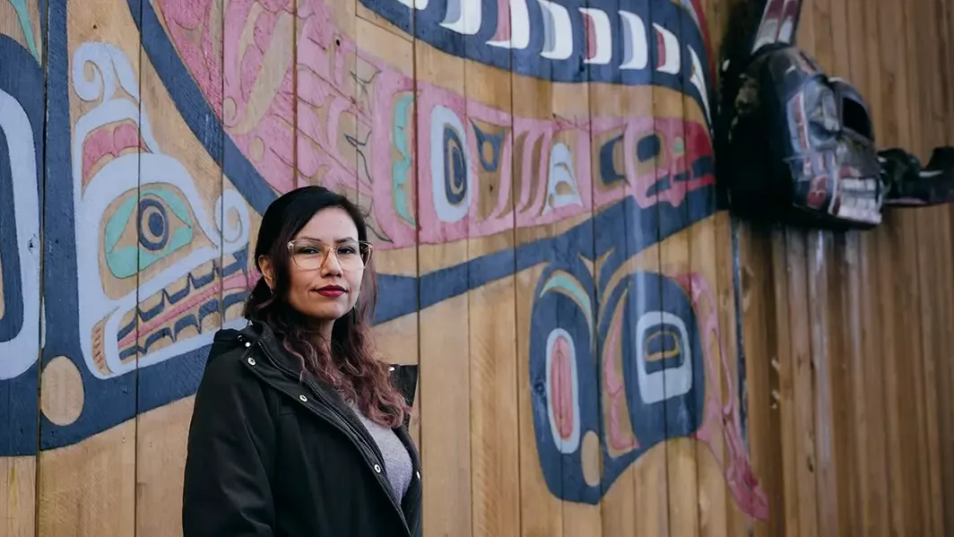 A woman wearing glasses and a dark coat stands beside a wooden wall featuring colourful Indigenous Northwest Coast-style artwork. Carved masks hang in the background as she looks toward the camera.