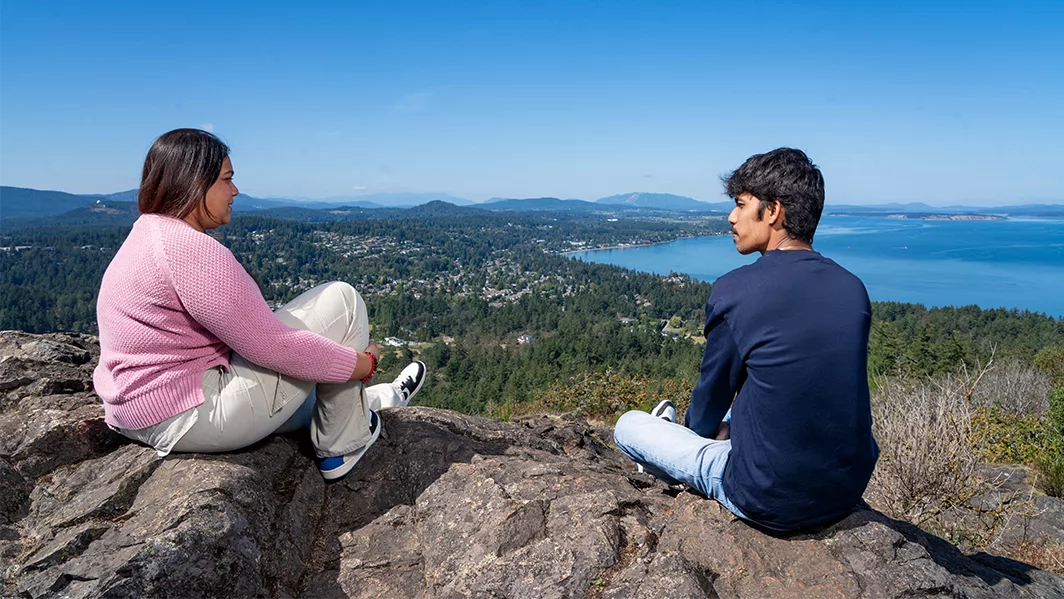Two students sit on a rocky hill that overlooks the Victoria landscape and ocean. 
