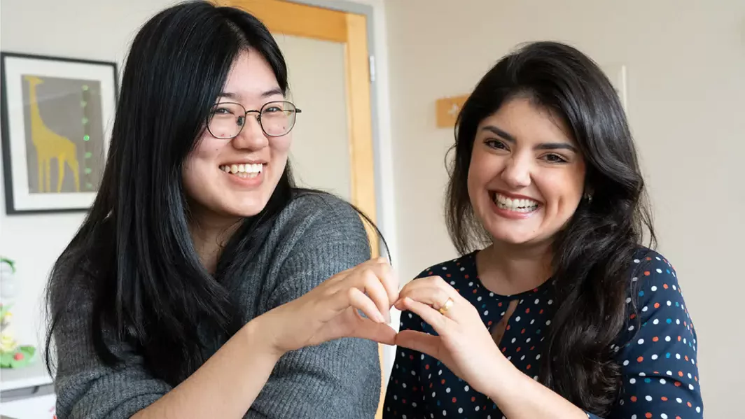 Two students smiling and making a heart sign with their hands. 