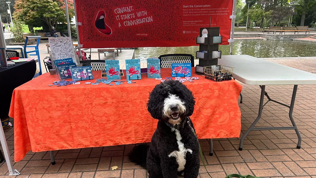 A black and white dog sits in front of an event table on campus.
