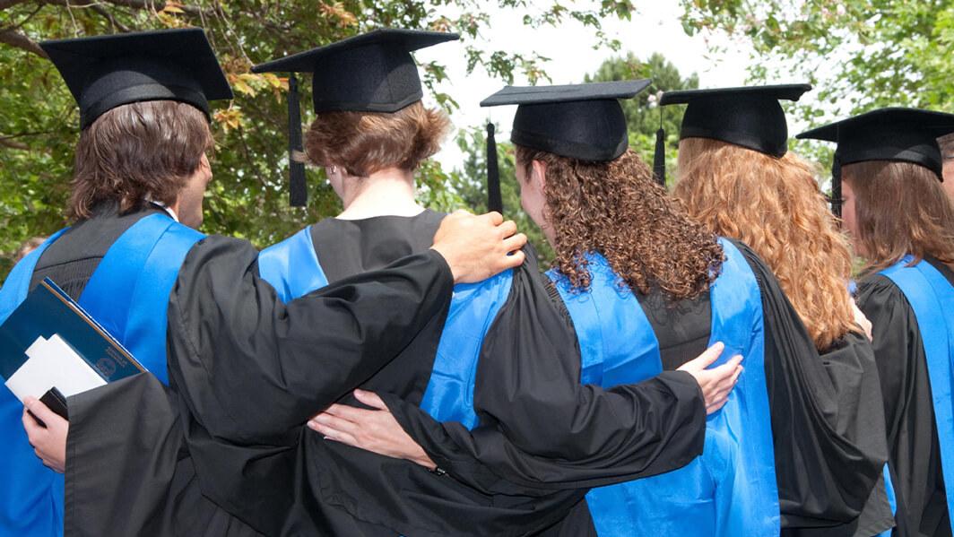 A group of students in their graduation regalia faces away from the camera, linking arms behind their backs