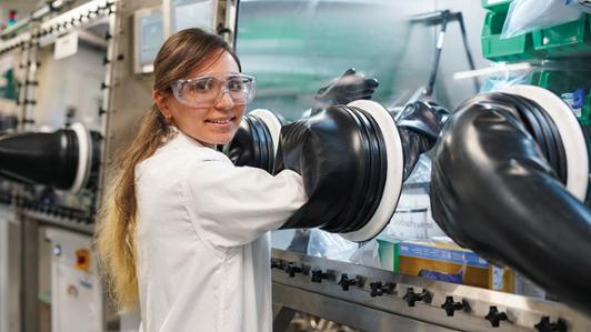 A female engineering graduate student wearing safety glasses and a lab coat smiles at the camera. She is in the Saidaminov Lab and her hands are in gloves in a clean, testing chamber in front of her.