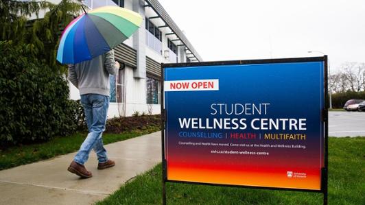 A person holding a rainbow umbrella walks towards the Student Wellness Centre. A sign says now open and lists counselling, health, and multifaith services.