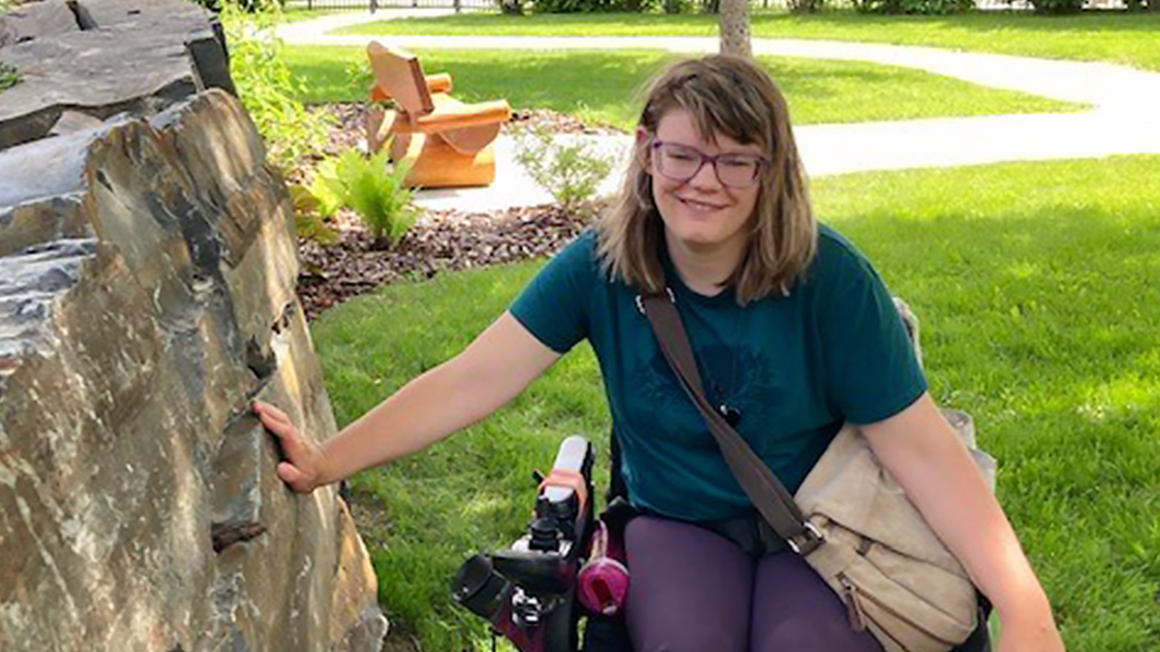 A woman in a wheelchair on a lawn smiles at the camera while touching a rock wall to her left