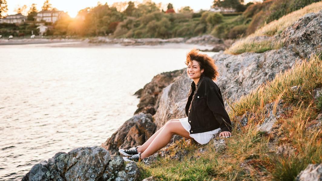 A woman with curly hair sits on rocks by the ocean smiling at the camera while the sunsets behind her.
