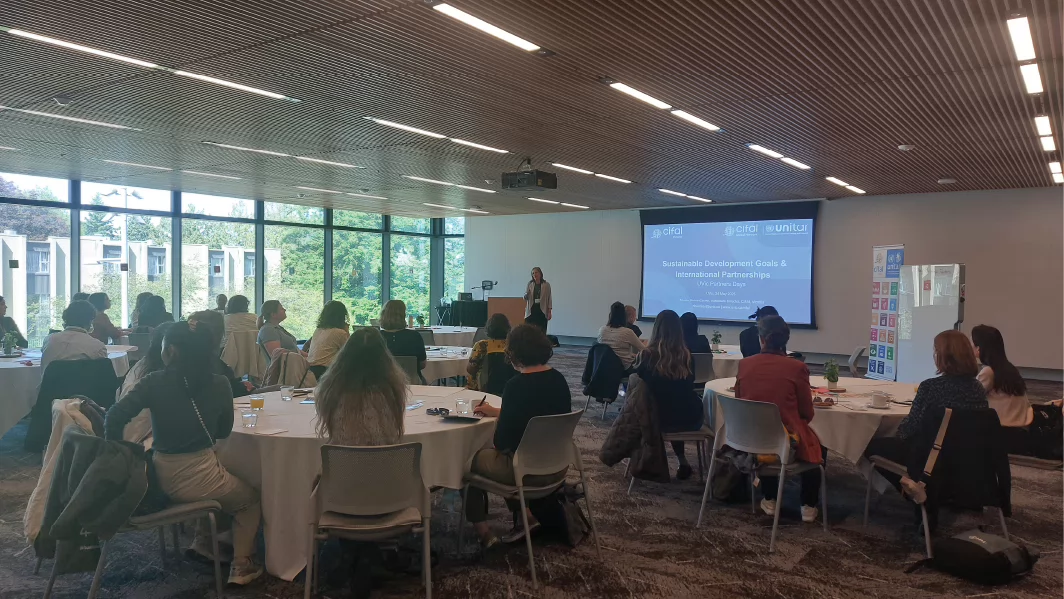Photo of a group of people in a conference room sitting in round tables while a speaker, Nicole Bates-Eamer, presents slides related to CIFAL Victoria and the UN SDGs
