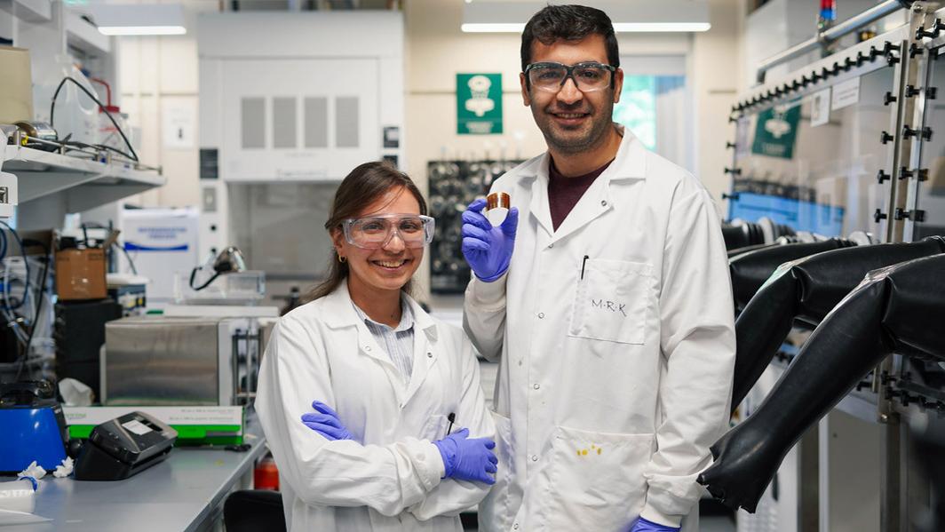 A female and male student in a lab and wearing white lab coats, safety goggles and purple gloves, smile at the camera. The man is holding a flexible solar cell between his thumb and index finger.