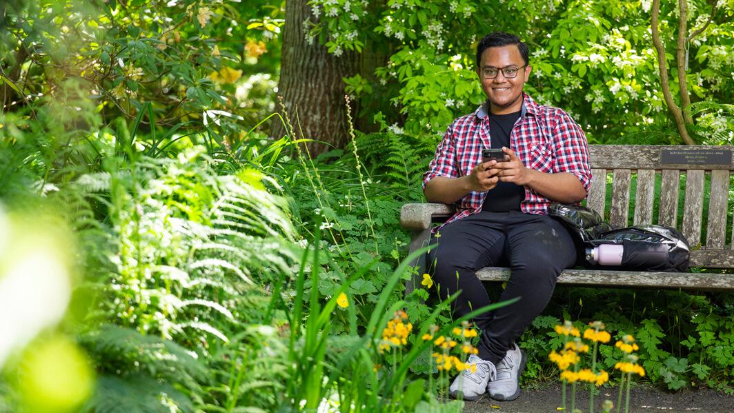 Shehnin Rahman smiles warmly at the camera as he holds his phone in both hands. He is sitting on a bench in a lush, green garden with yellow flowers.