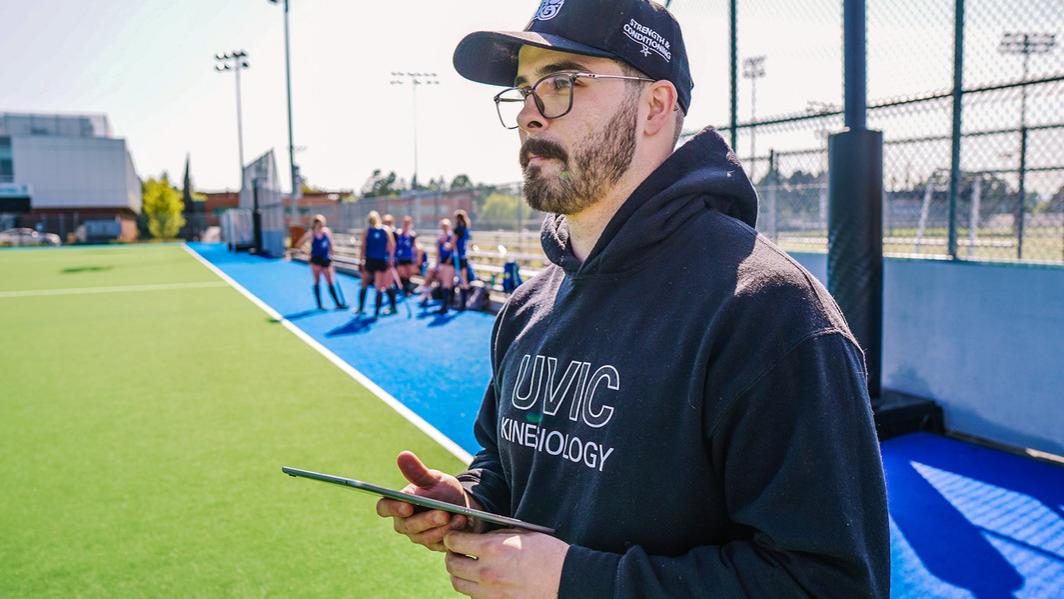 A male student wearing a black UVic Kinesiology hoodie and Vikes "strength and conditioning" hat holds an iPad and looks off camera onto the field hockey turf. Members of the womens field hockey team are out of focus in the background behind him.