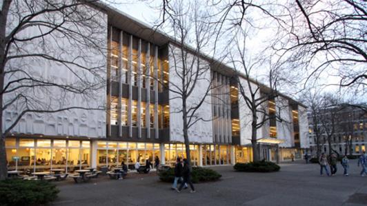 Photo of the exterior at dawn of UVic's McPherson Library with the interior lights on and some people walking by 