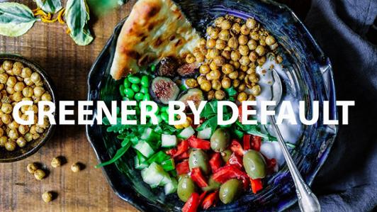 Photo of two handcrafted bowls on top of a wooden board filled with of tasty vegetarian food (green beans, olives, red pepper, figs, bread, etcetera), with the legend: "Greener by default" 