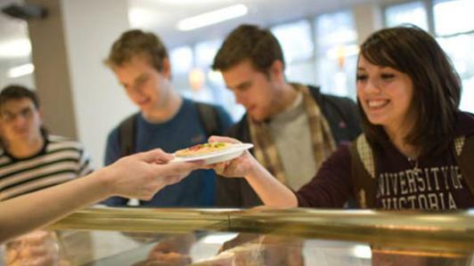 Photo of a UVic dining outlet serving food to smiling students. The young female student receiving a plate from the server wears a University of Victoria sweatshirt