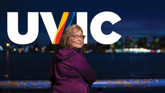 Photo of UVic's health researcher Bernie Pauly holding a hand rail in an ocean font at dusk, looking back at the camera with the blurry lights of a city in the background across the sea. The logo of UVIC is positioned behind her.