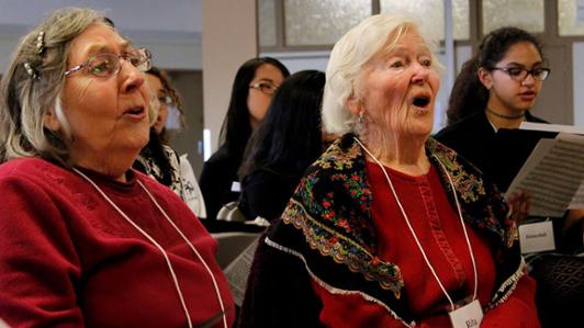 Image of an intergenerational choir performing, showing two senior ladies in the front row, and multiple young female singers in the background