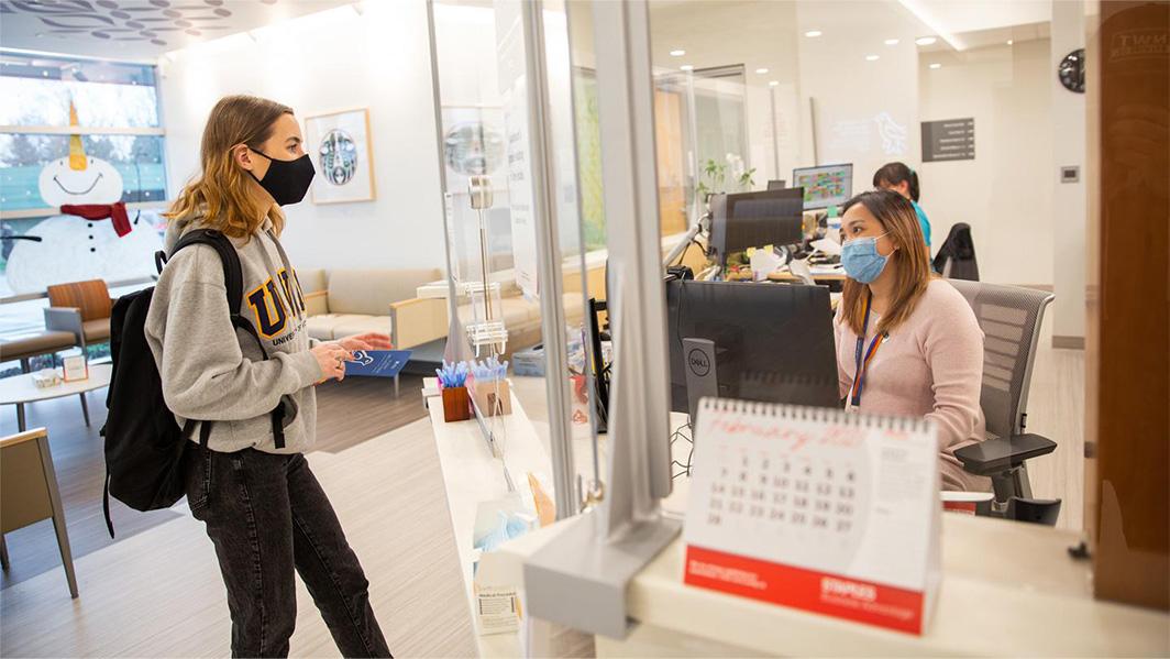 Photo of a young female student interacting with staff at the front desk of the Student Wellness Centre at UVic.
