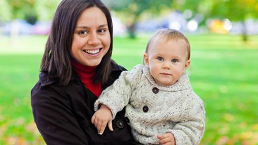 Photo of a smiling woman carrying a toddler with a green park in the background