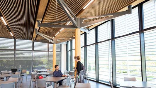 Photo of the interior of UVic's new housing building, Cheko’nien House, with glass walls and wooden ceiling, showing two students interacting 