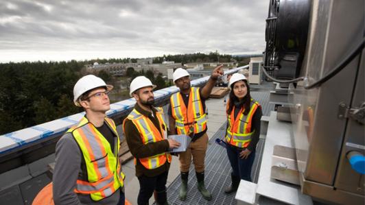 Photo of Ralph Evins, Haris Shamsi, Rajeev Kotha and Mahsa Torabi wearing safety gear (vest and helmets) while inspecting an AC system on top of a UVic building 