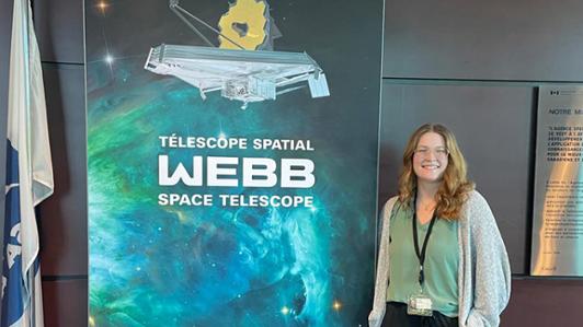 Photo of the student Tess Grindlay, in front of a poster of the James Webb Space Telescope at the Canadian Space Agency 
