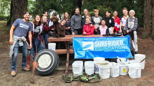 Group photo of sixteen young volunteers (and a furry dog) behind  bins containing the waste collected from the campaing. They hold a sing of the "Surfrider Foundation"
