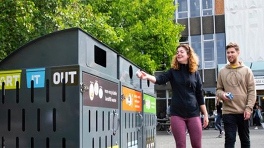 Photo of a female and male UVic staff members disposing waste into the large waste bins distributed on campus with colour signages for each type of waste