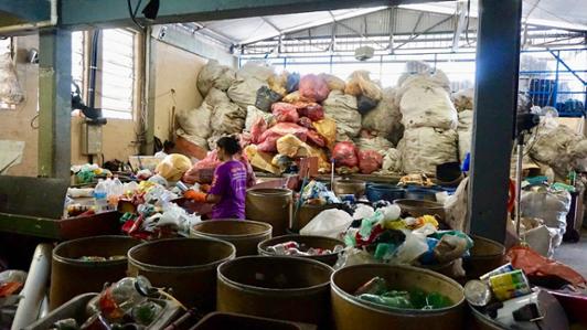 Photo of a waste recycling centre from the Cooperativa Cooperfenix - Centro de Reciclagem, at Diadema, Brazil, showing the inside of a warehouse with tons of different types of waste separated by type  in large sacks and metal cans , with a woman working in the middle with gloves.
