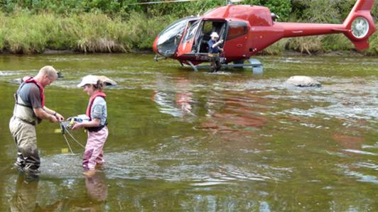 Photo of a stationary helicopter on the water  while two researchers take water samples from the Ells River in Northern Alberta 