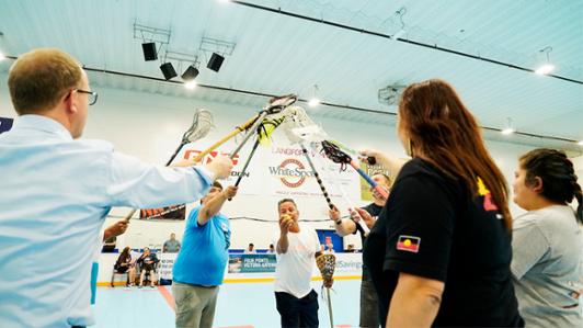Photo of seven representatives of different indigenous nations from the Commonwealth holding their Lacrosse sticks towards the centre, as  part of an exhibition lacrosse game.
