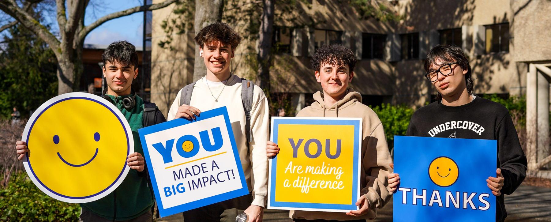 Four male students stand smiling at the camera, one holds a yellow smiley face sign and the three others hold blue and yellow thank you signs 