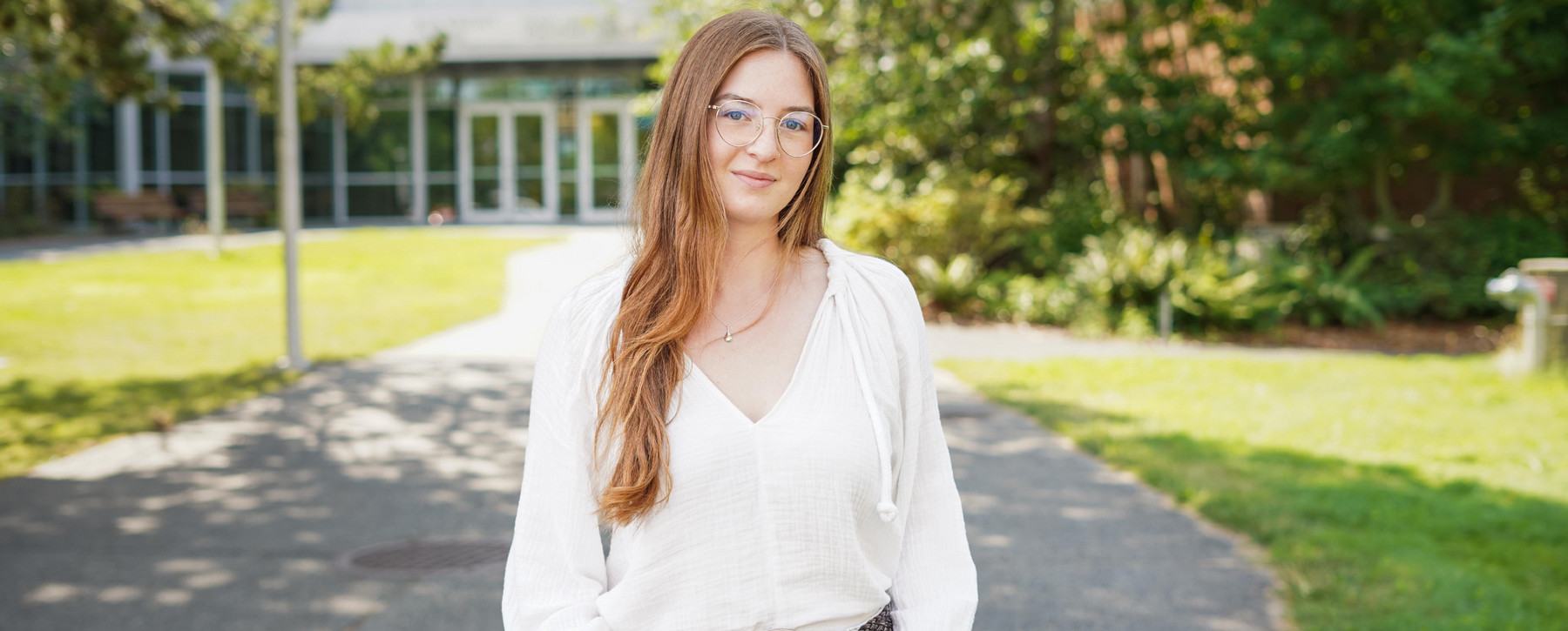 A student with long hair and glasses in a white blouse stands on the path leading to the UVic Bob Wright building