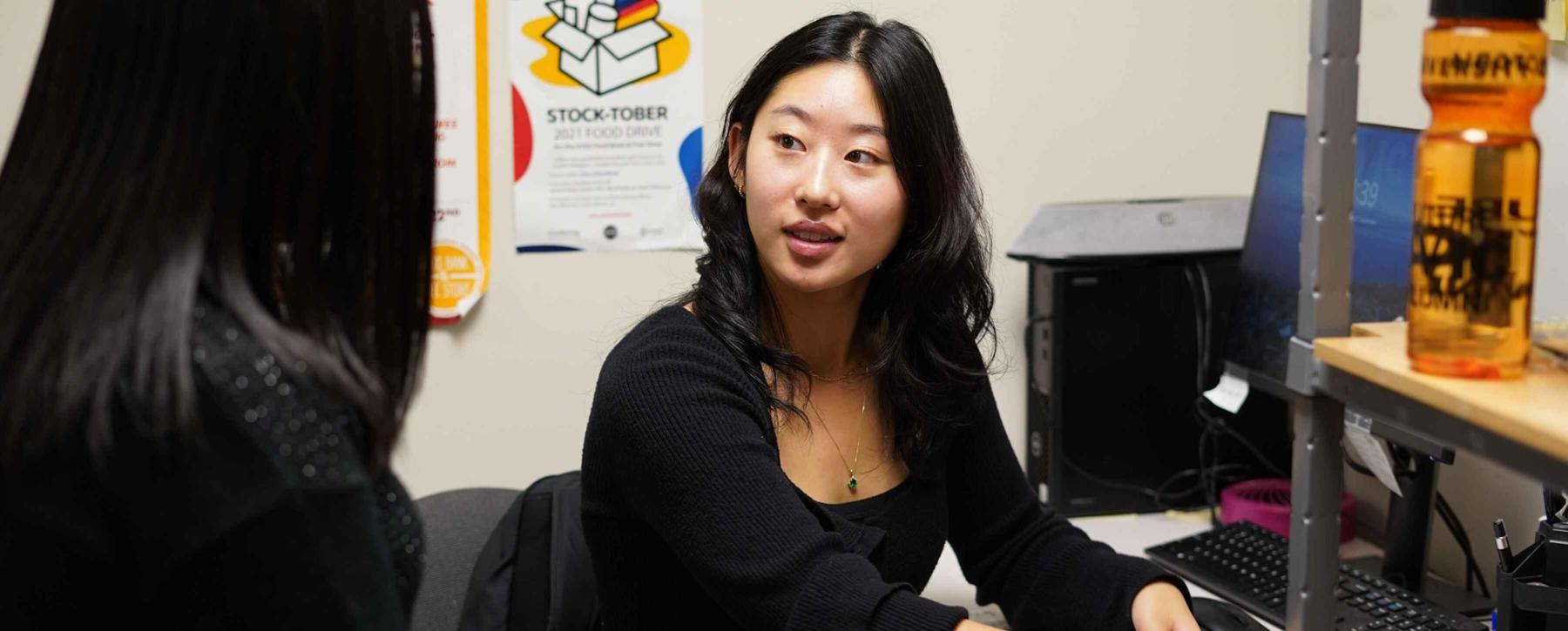 Crystal Ni sits at the desk in the food bank talking to a woman with her back to the camera