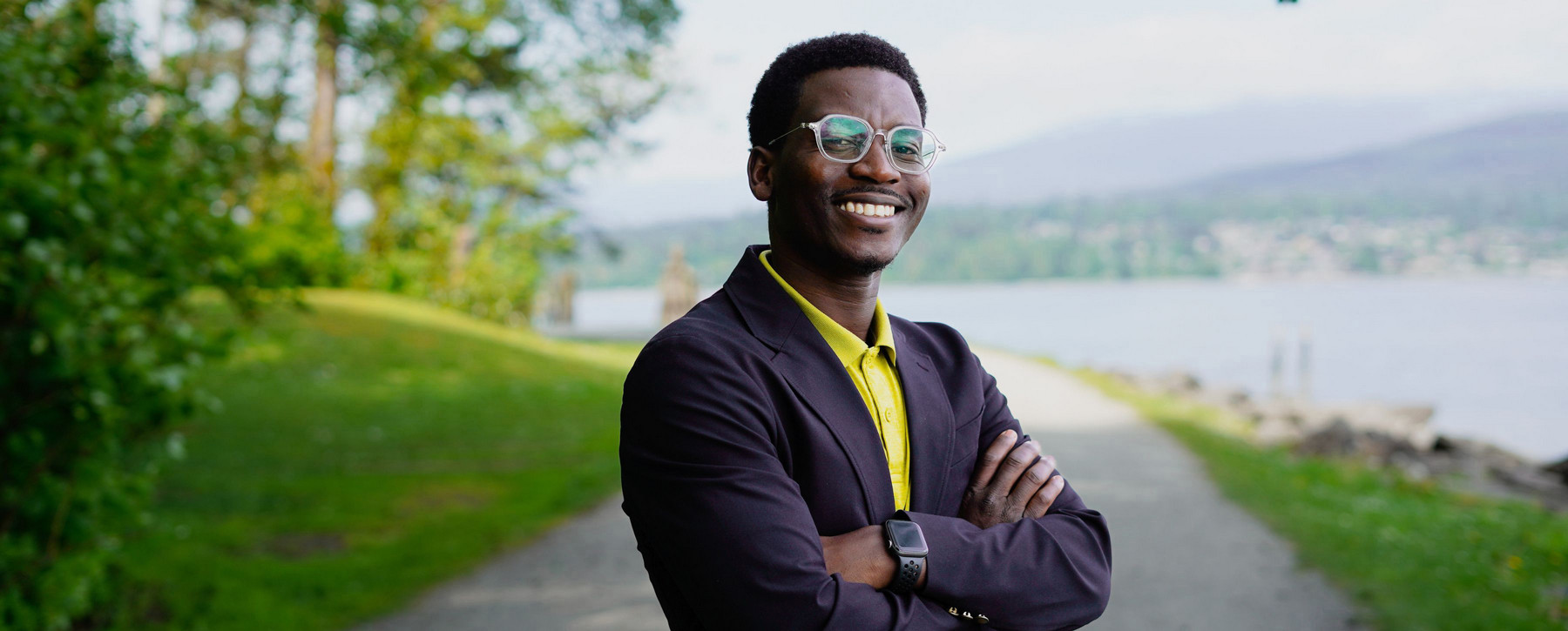 A person standing on a path, arms folded, but smiling, surrounded by greenery with the ocean in the background.