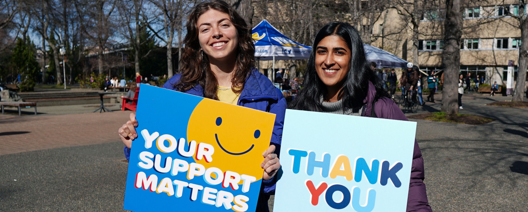 Two students in quad holding signs that say "Your support matters" and "thank you"