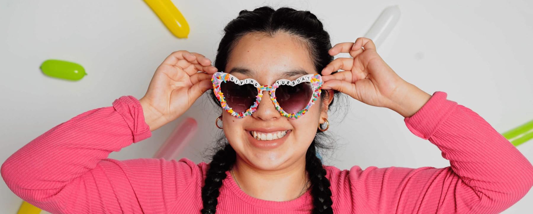 A student with two braids in a pink sweater is wearing sprinkle glasses that say "Giving Tuesday" in beads on them. She's standing in front of a white background with colored sprinkle balloons.