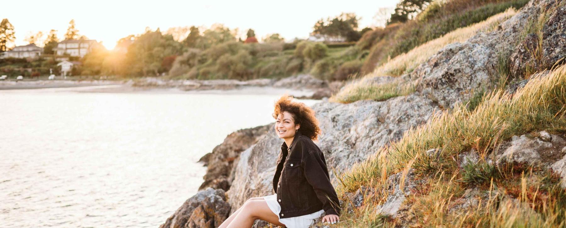 A woman with curly hair sits on rocks by the ocean smiling at the camera while the sunsets behind her.
