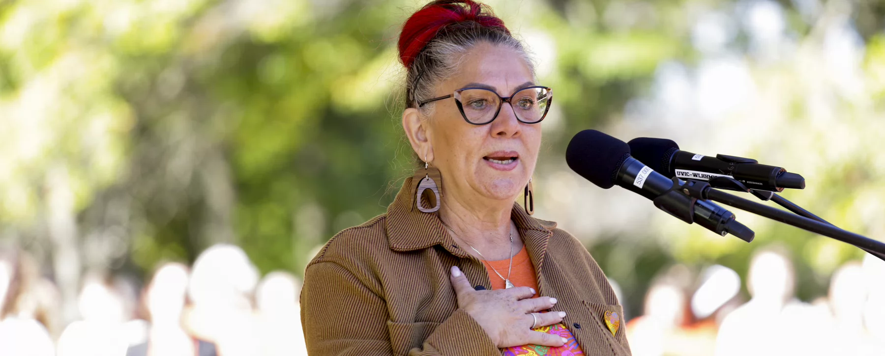Qwul’sih’yah’maht, Robina Thomas stands at a microphone wearing an orange shirt and placing her hand on her heart.