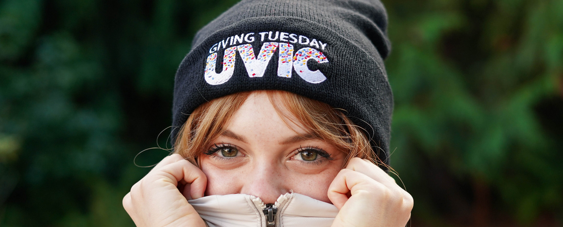 A person peeking out of zipped up coat wearing a black toque embroidered with Giving Tuesday