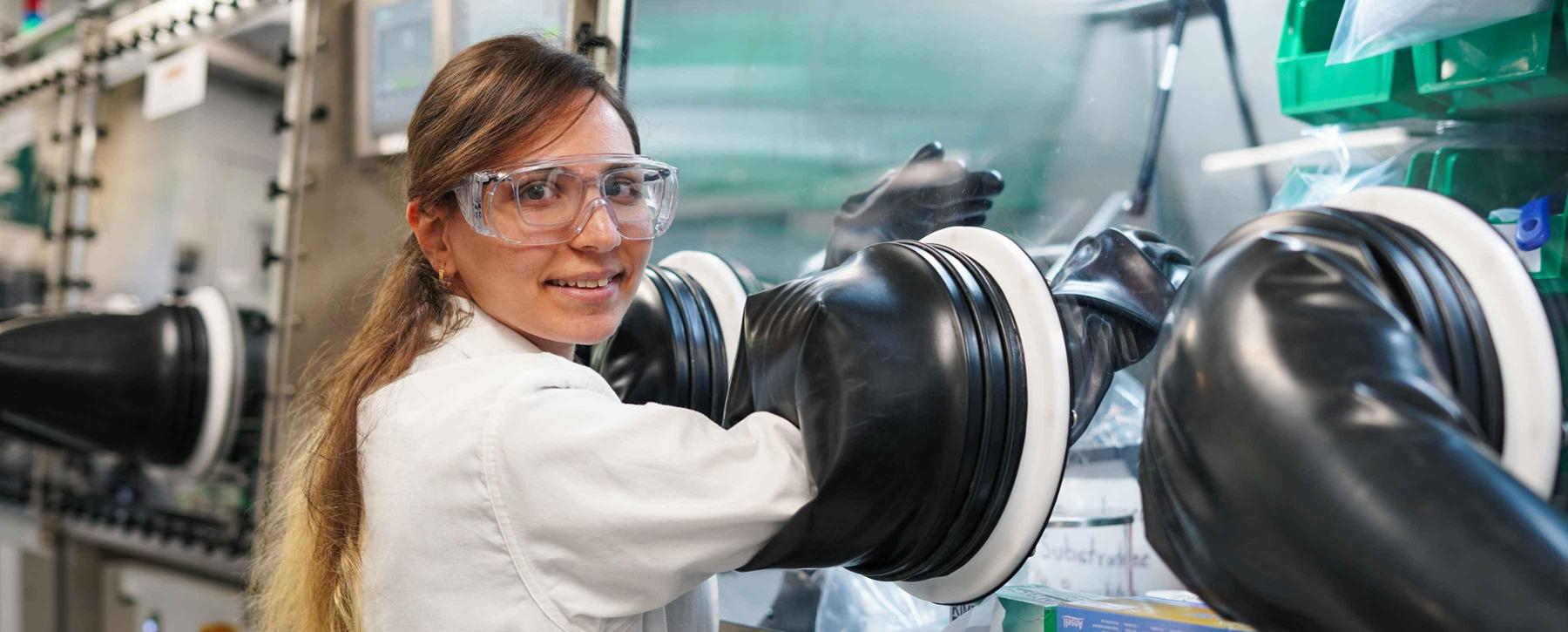 A female student wearing safety glasses and a lab coat smiles at the camera. She is in a lab and her hands are in a chamber in front of her.