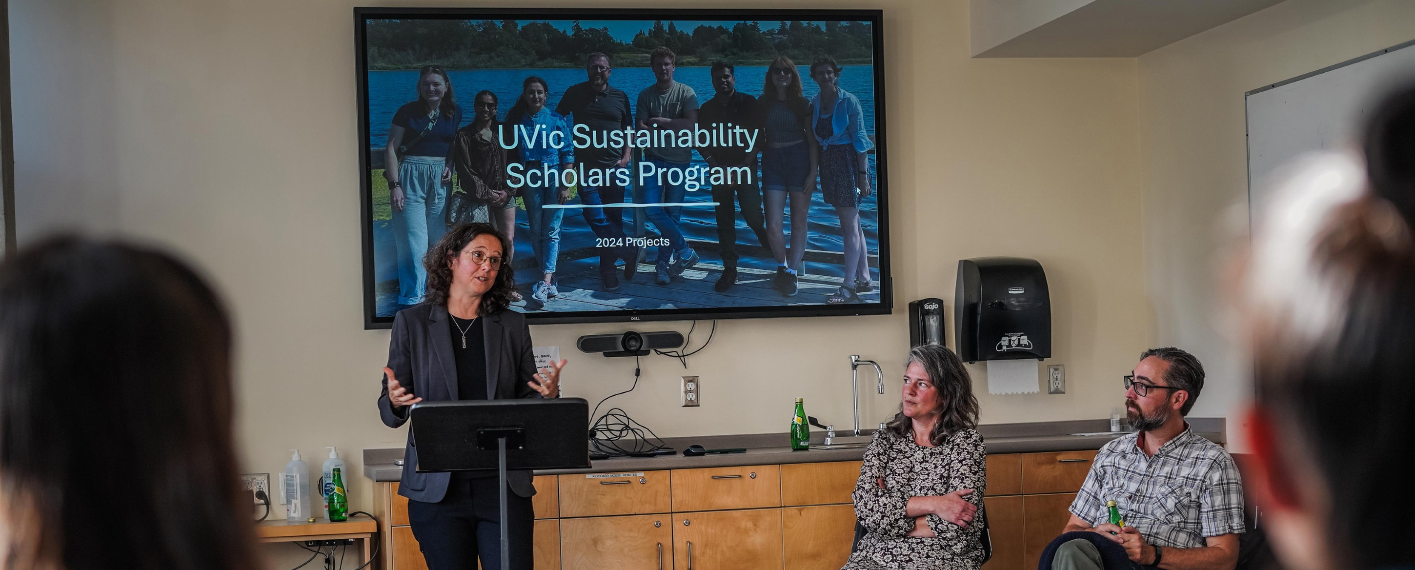 A woman giving a presentation to a classroom with 'Sustainability Scholars Program' displayed on a screen behind her.