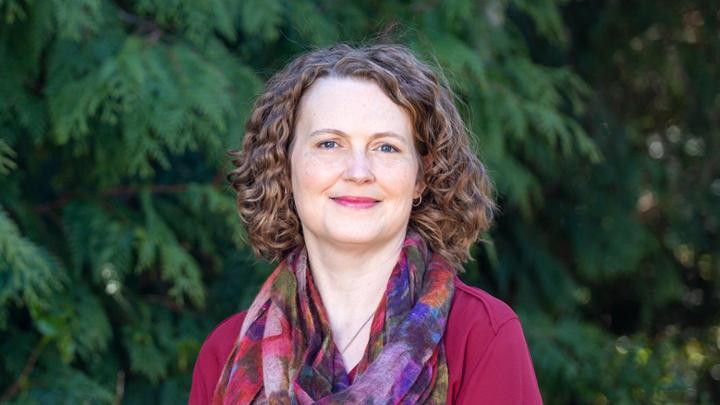 Photo of Christina Harris standing in front of blurry cedar trees. She is a white woman with curly brown hair, wearing a red shirt and an colourful scarf.