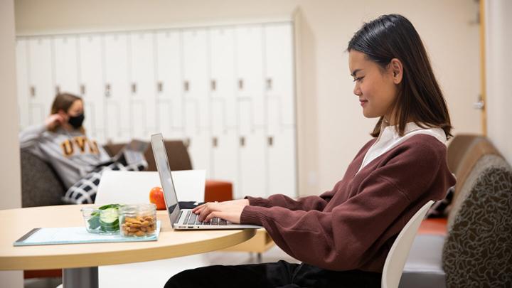 A student sits at a table eating lunch in front of their laptop. Another student is wearing a mask in the background reading a book.