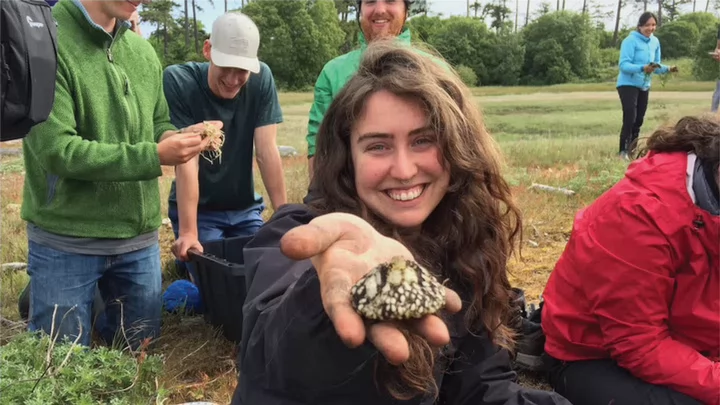 Photo of a UVic student smiling to the camera while holding a mushroom, as part of the Students at the Tl’ches Ethnoecology Field School