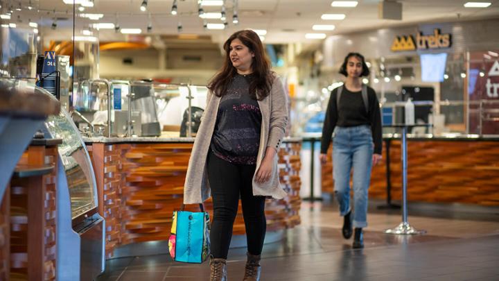 Two women walk through Mystic Market. The woman in foreground looks at a display of baked goods at a coffee shop.